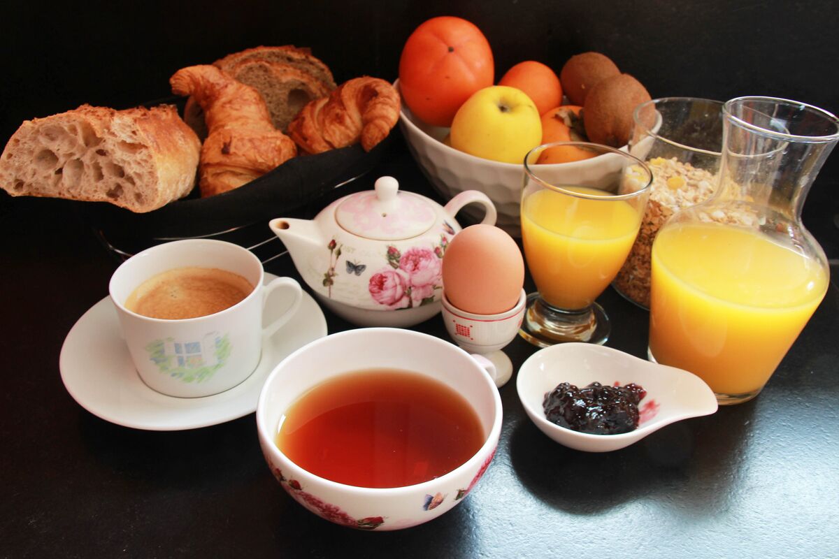 A breakfast setting with croissants, a teapot, a cup of tea, a bowl of fruit, and a glass of orange juice.