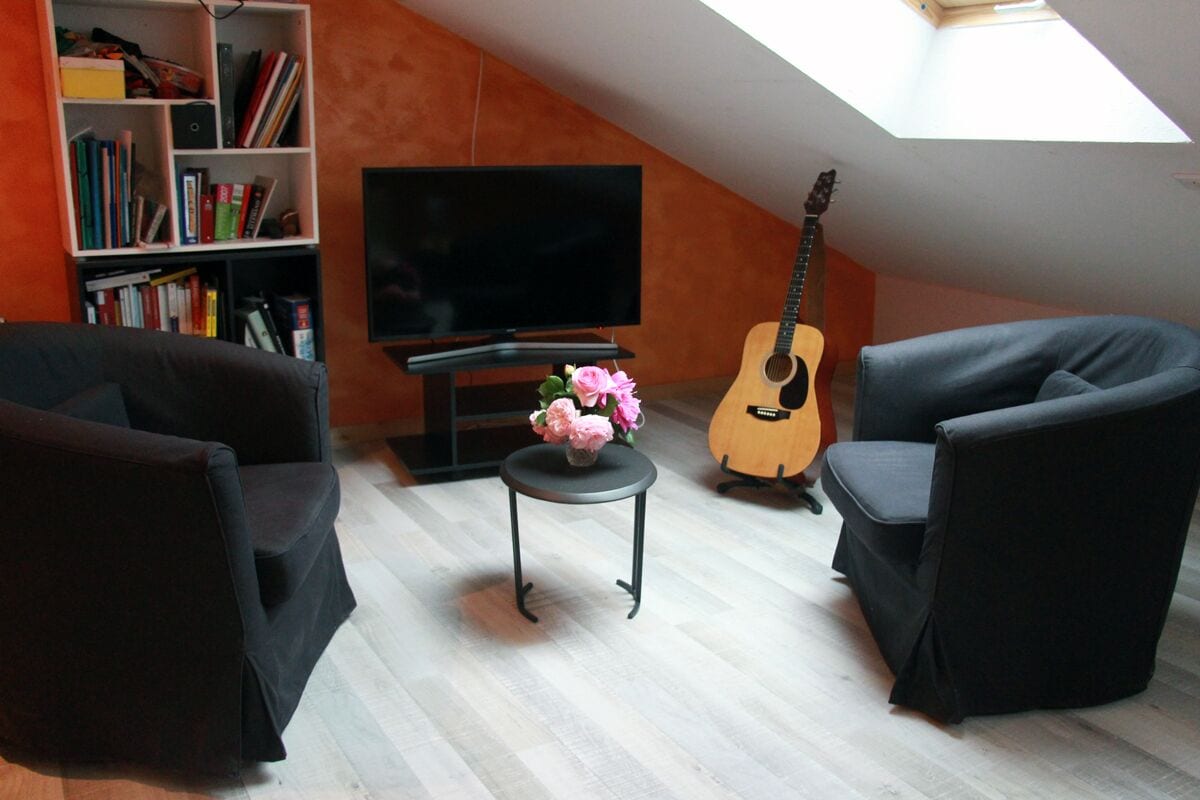 A living room with a flat-screen TV, a guitar, a bookshelf, and a small table with flowers.