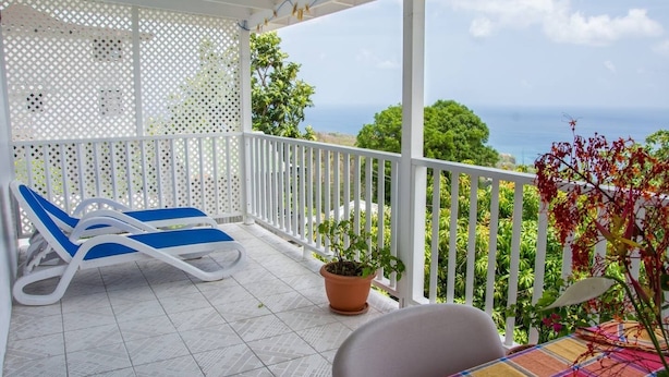 A balcony with a white railing, a blue lounge chair, a potted plant, and a table with a colorful tablecloth.