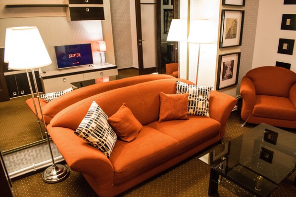 A modern living room with an orange sofa, a glass coffee table, and a flat-screen TV on a stand.