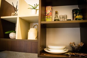 A kitchen area with shelves containing various items including a white pitcher, plates, and a bottle of Jacobs Krönung beer.
