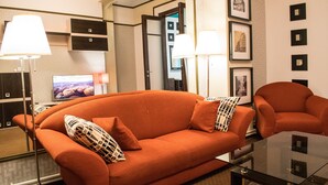 A modern living room with a large orange sofa, two side tables with lamps, a flat-screen TV, and framed pictures on the wall. - ❤️ CityCenter Athenaeum Exquisite 1BD Apartment ❤️ (Sector 1)