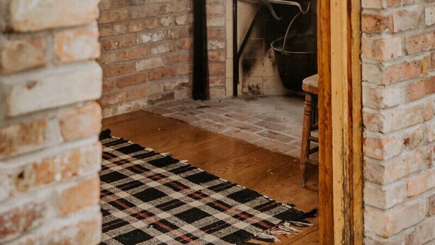 Un intérieur rustique avec des murs en briques, une cheminée, un tabouret en bois et un tapis à carreaux.