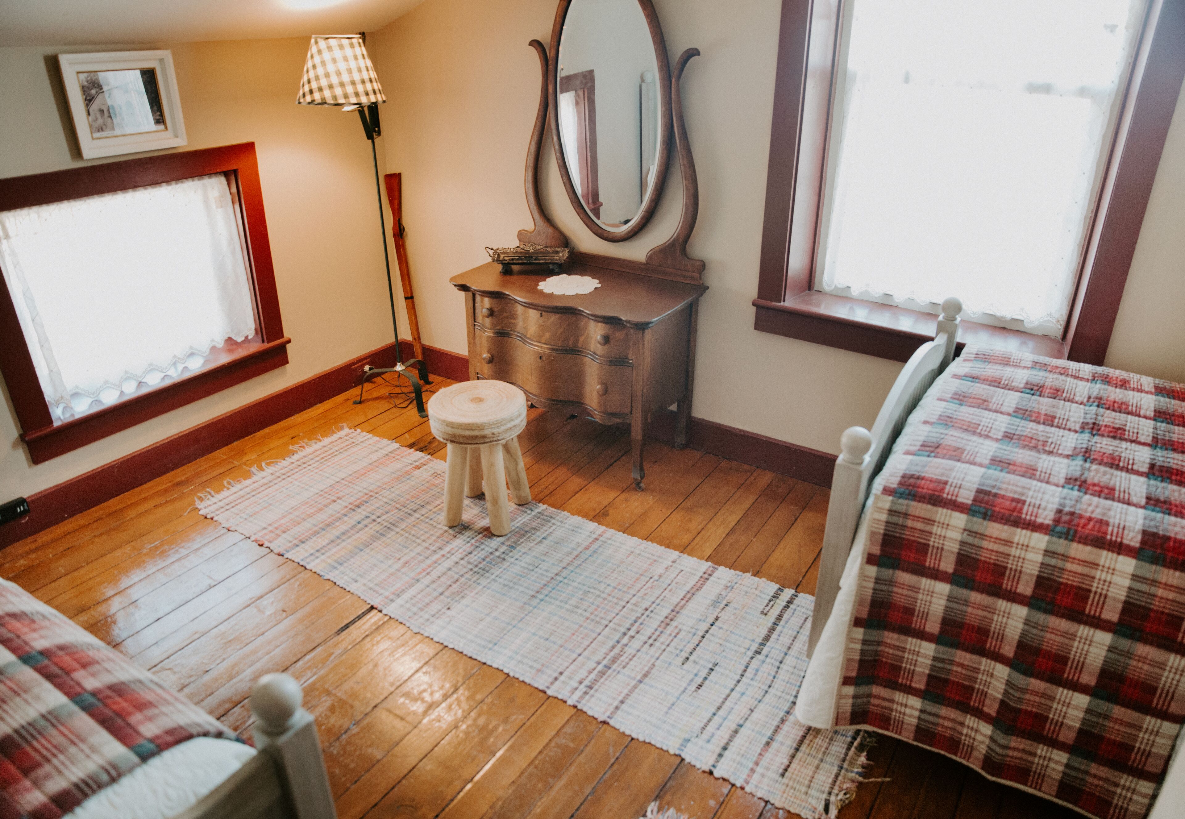 Une chambre avec un plancher en bois, un lit recouvert d’une couverture à carreaux, une commode avec un miroir et une fenêtre donnant sur un paysage agréable.