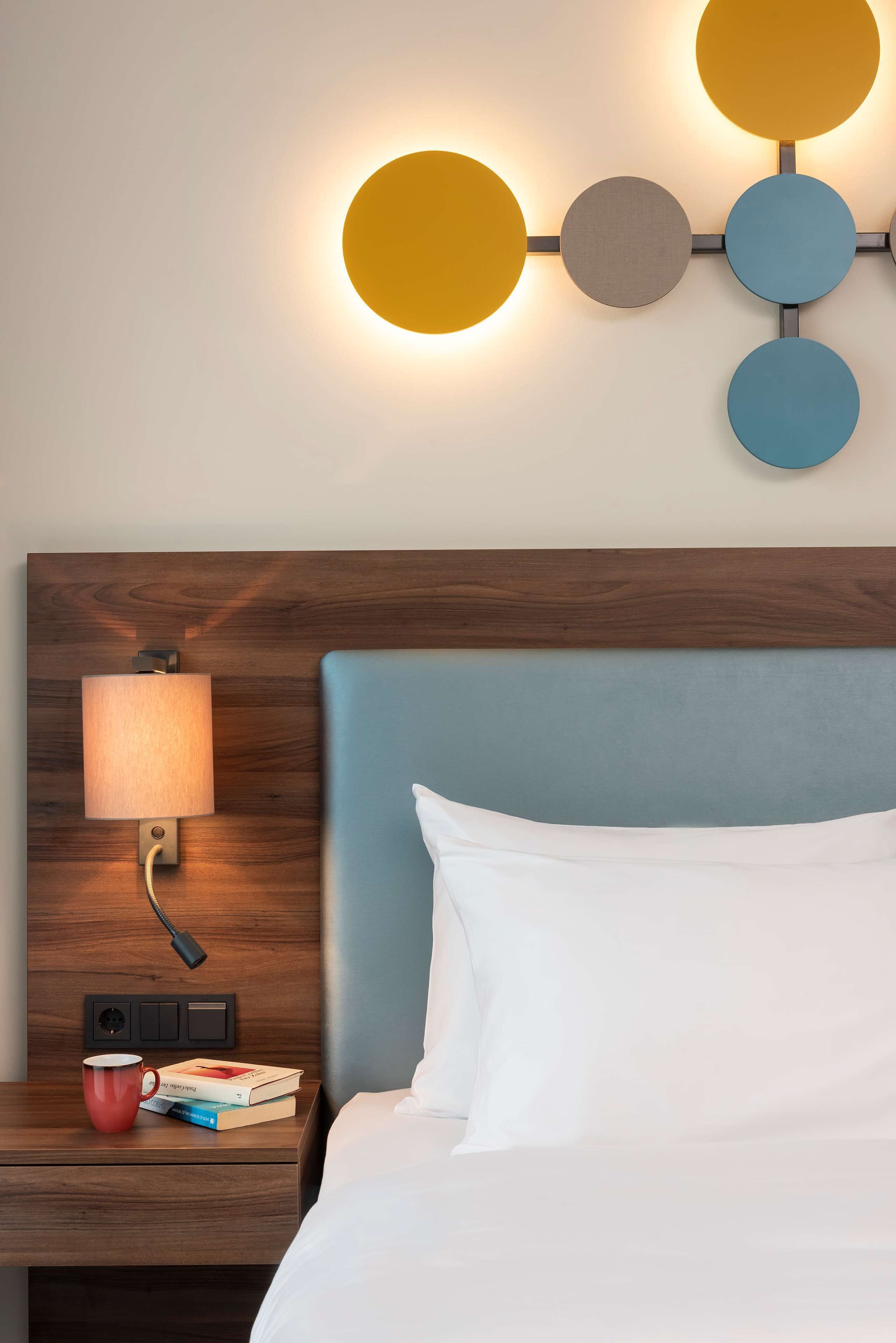 Una habitación de hotel con cabecera de madera, una lámpara de noche, una taza roja y un libro sobre la mesita de noche.