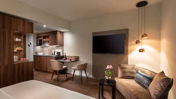 Una habitación de hotel moderna con una cama, un sofá, una pequeña mesa con un florero, un televisor montado en la pared y una cocina al fondo.