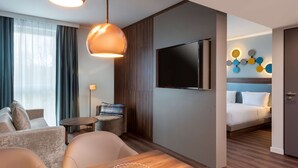 Une chambre d’hôtel moderne équipée d’un canapé, d’un fauteuil, d’une table basse en bois et d’un téléviseur fixé au mur.