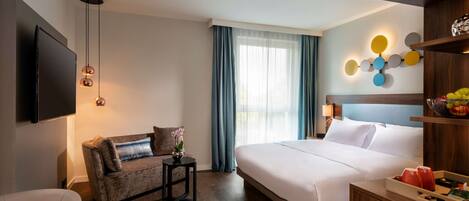 Habitación de hotel con cama, sofá, televisor, mesita y lámpara de pared.