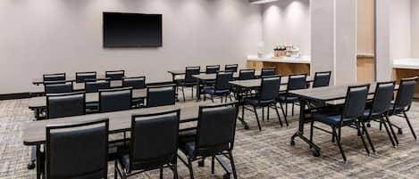 A conference room with multiple tables and chairs, a flat-screen TV mounted on the wall, and a kitchenette area.