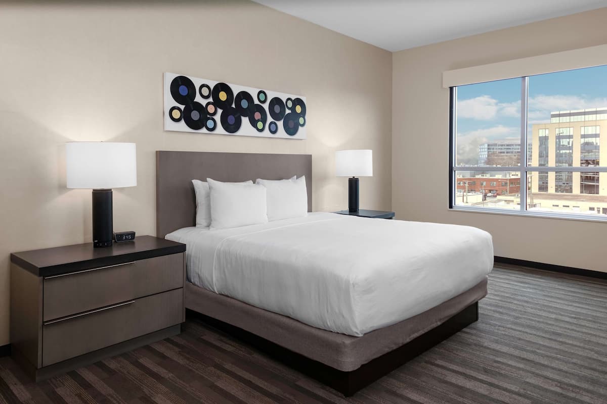 Ein modernes Hotelzimmer mit einem großen Bett, zwei Nachttischen, einem Wandbild und einem großen Fenster mit Blick auf die Stadt.