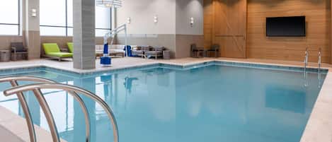 Una piscina interior moderna con agua azul cristalina, un área de descanso junto a la piscina con sillas verdes, y una pared de madera con un televisor montado.