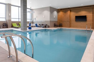 Una piscina interior moderna con agua azul cristalina, un área de descanso junto a la piscina con sillas verdes, y una pared de madera con un televisor montado.