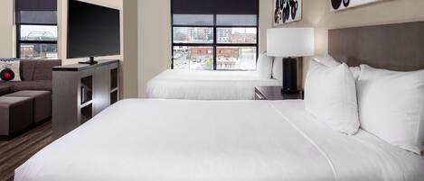 Ein Hotelzimmer mit einem großen Bett, einem Fernseher, einem Schreibtisch und einem Sofa. Ein Fenster bietet Blick auf die Stadt.