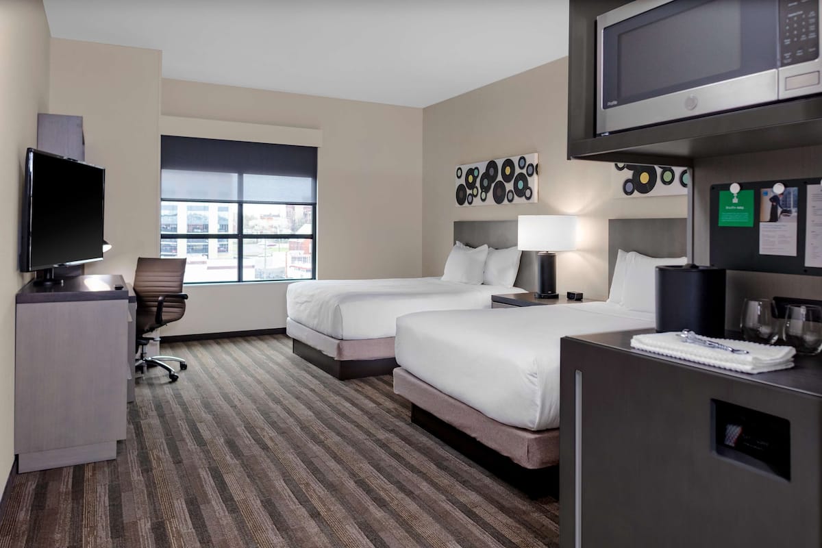 Ein Hotelzimmer mit zwei Betten, einem Schreibtisch, einem Fernseher, einer Mikrowelle und einem Fenster mit Blick auf die Stadt.