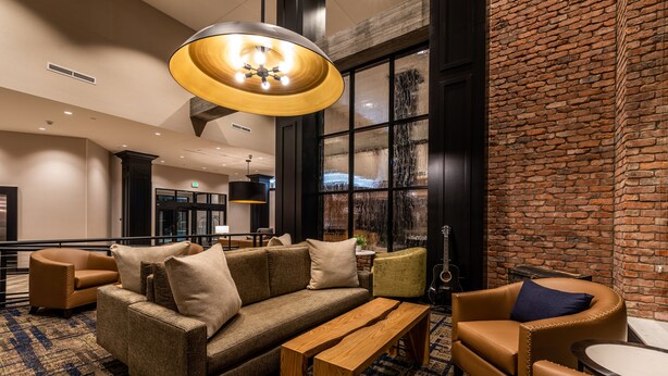 A lobby with a brown sofa, a wooden bench, and a round table. There is a brick wall and a large window with a view. - Hyatt House Nashville Downtown/Convention Center (Nashville)