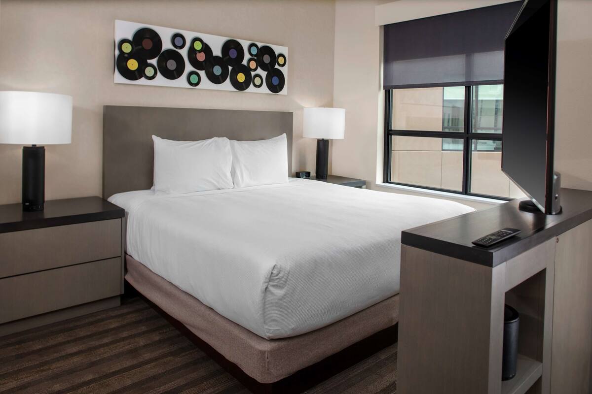 Ein Hotelzimmer mit einem großen Bett, zwei Nachttischen mit Lampen, einem Schreibtisch mit Computer und einem Fenster mit Blick.