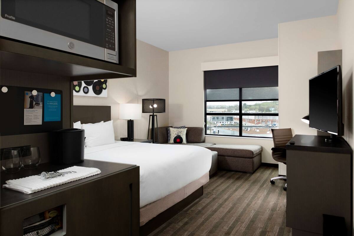 Ein Hotelzimmer mit einem großen Bett, einem Schreibtisch mit Computer, einem Fernseher, einem Fenster mit Stadtblick und einer Minibar.