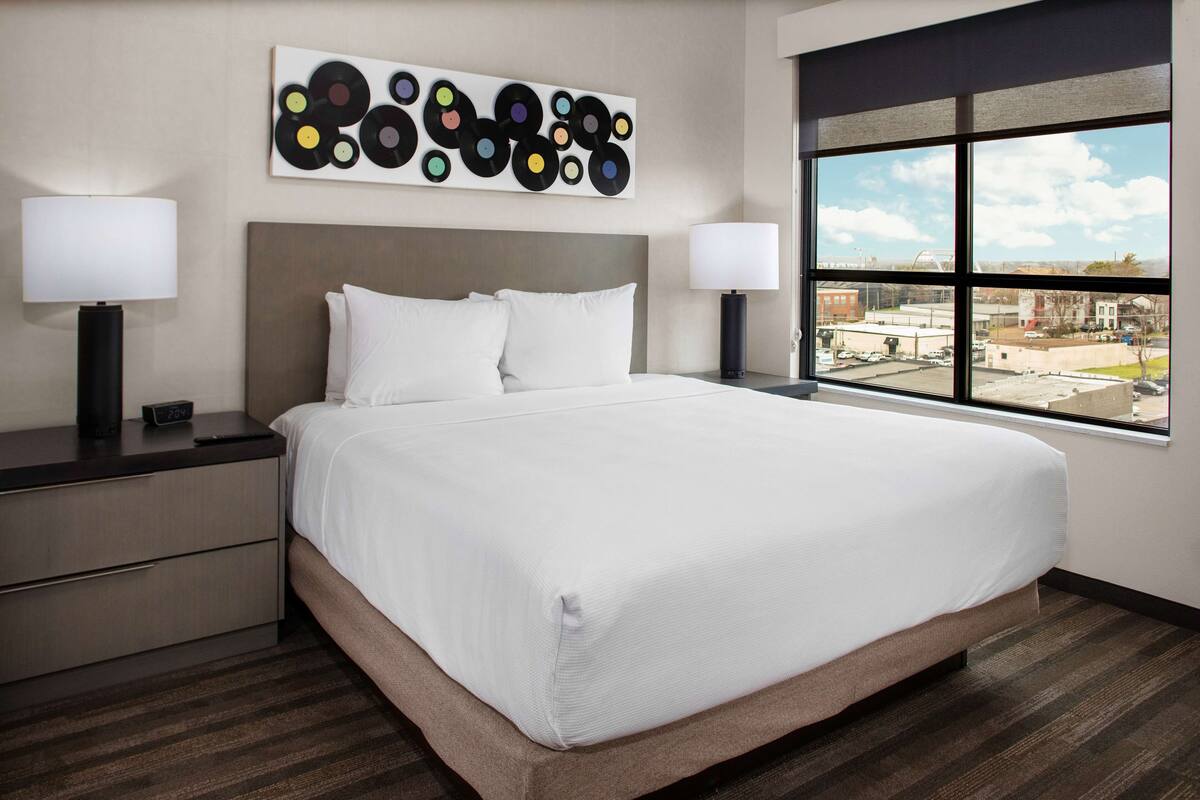 Ein Hotelzimmer mit einem großen Bett, zwei Nachttischen mit Lampen, einem modernen abstrakten Wandbild und einem großen Fenster mit Blick auf die Stadt.