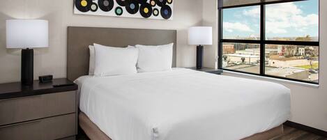 Habitación de hotel con una cama grande, dos mesitas de noche con lámparas, una obra de arte abstracta moderna en la pared y un amplio ventanal con vistas a la ciudad.