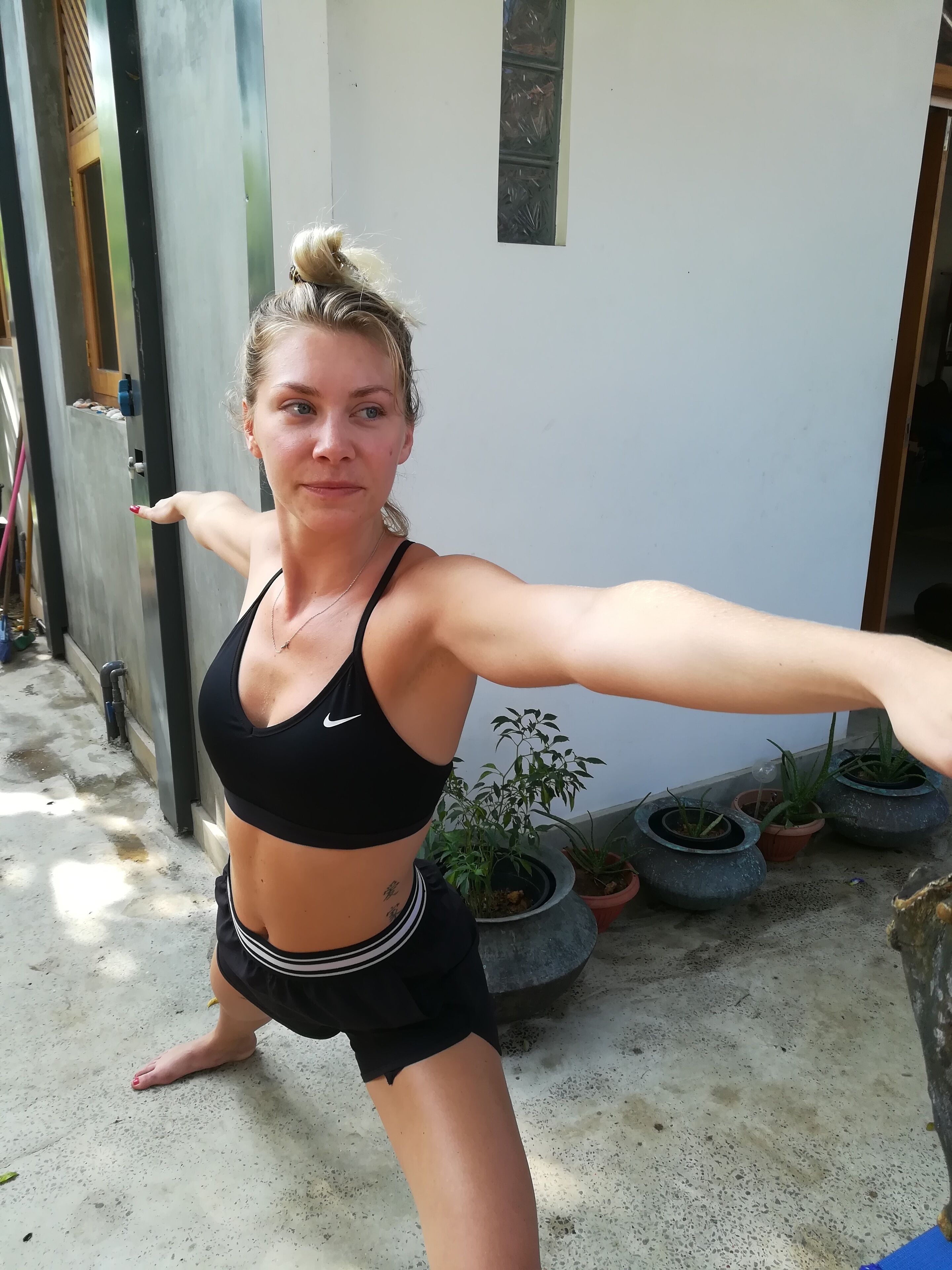 Une personne vêtue d’une tenue de sport noire, debout à l’extérieur, avec des plantes en pot et un sol en béton.