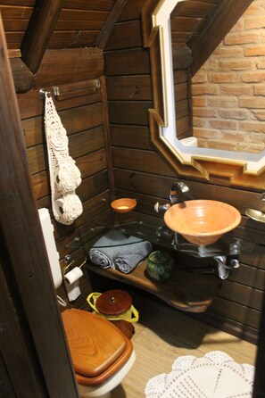 Un baño pequeño con paredes y retrete de madera, un lavabo con cuenco, y un estante con un cuenco y una botella verde.