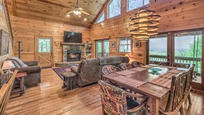 A spacious cabin with a large living area, a stone fireplace, and a dining table with rustic chairs. - Adventure Us- Aska Area, Lake Views, Dog Friendly (Blue Ridge)