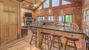 A wooden cabin interior with a stone fireplace, a bar area with stools, and large windows. - Adventure Us- Aska Area, Lake Views, Dog Friendly (Blue Ridge)