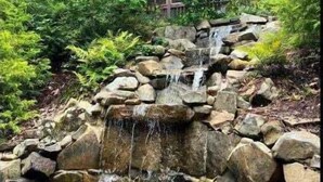 A waterfall cascading over rocks in a lush garden setting with dense foliage and a building in the background. - Adventure Us- Aska Area, Lake Views, Dog Friendly (Blue Ridge)