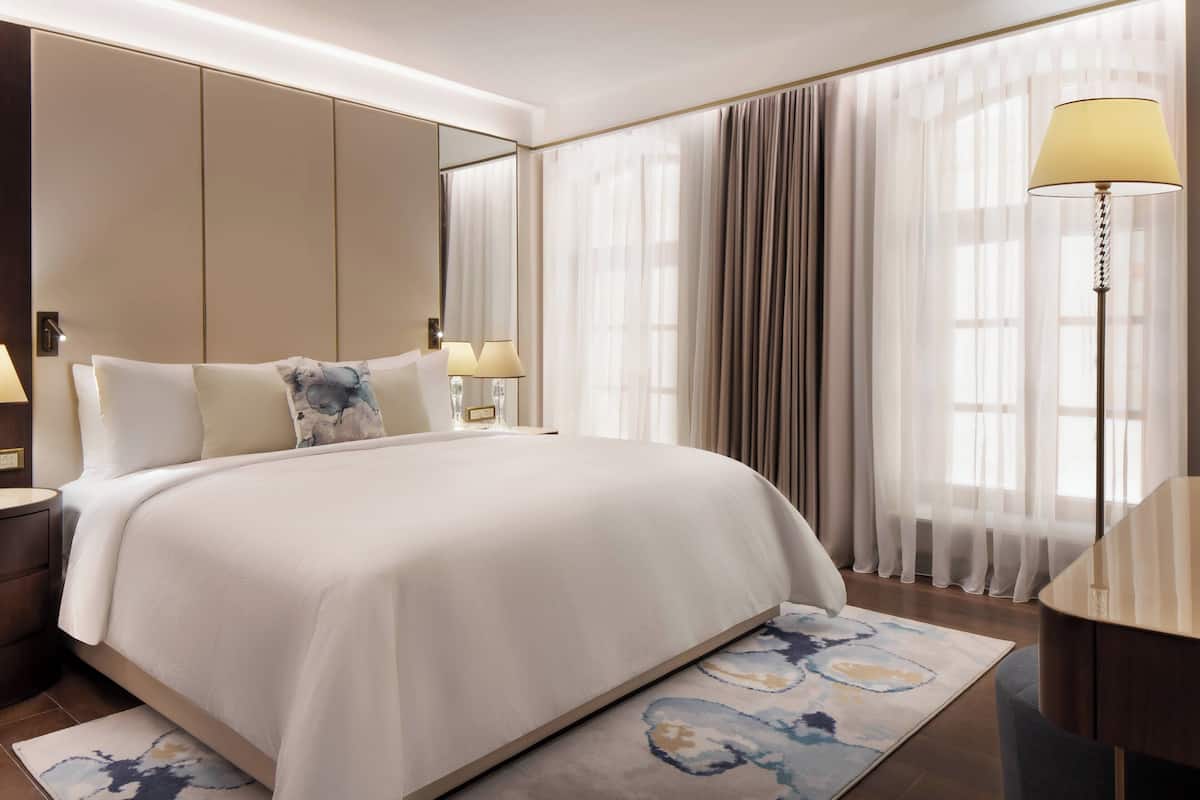 Habitación de hotel moderna con una cama grande, una mesita de noche con lámparas, un espejo y vistas a la ciudad a través de cortinas translúcidas.