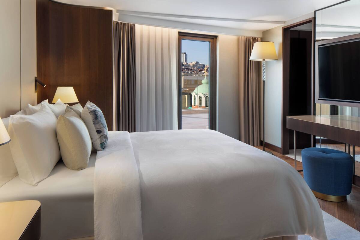 Habitación de hotel con una cama grande, un escritorio, un televisor y vistas de la ciudad a través de una ventana.