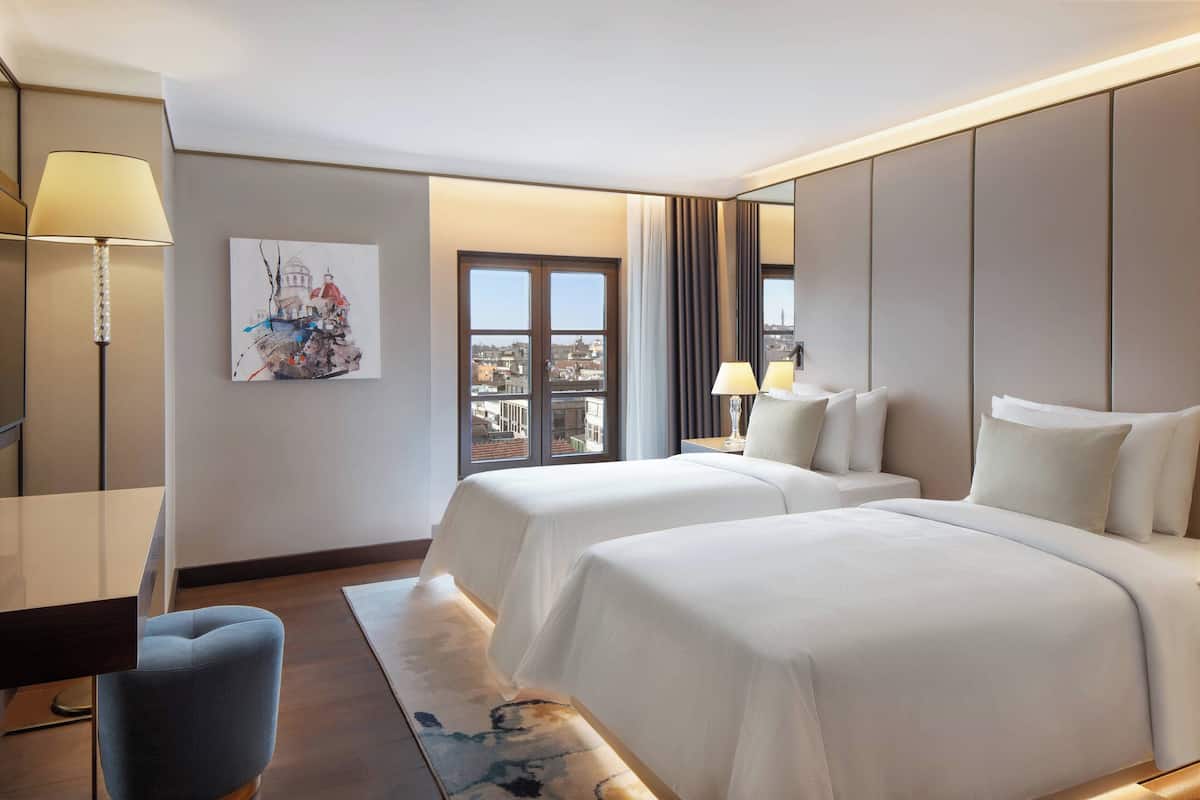 Habitación de hotel con dos camas, un escritorio, una silla, una lámpara y un cuadro en la pared.