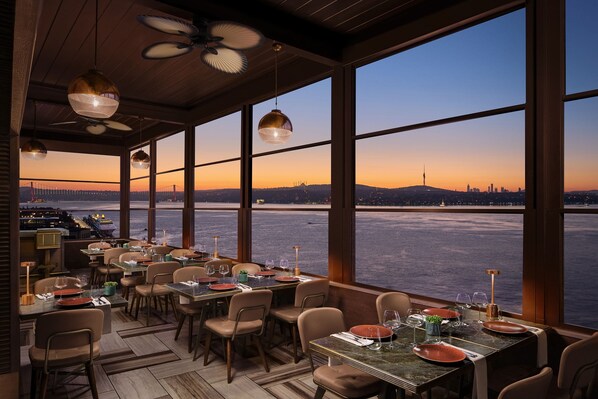 A restaurant with tables set for dining, large windows with a view of a city skyline and water body.