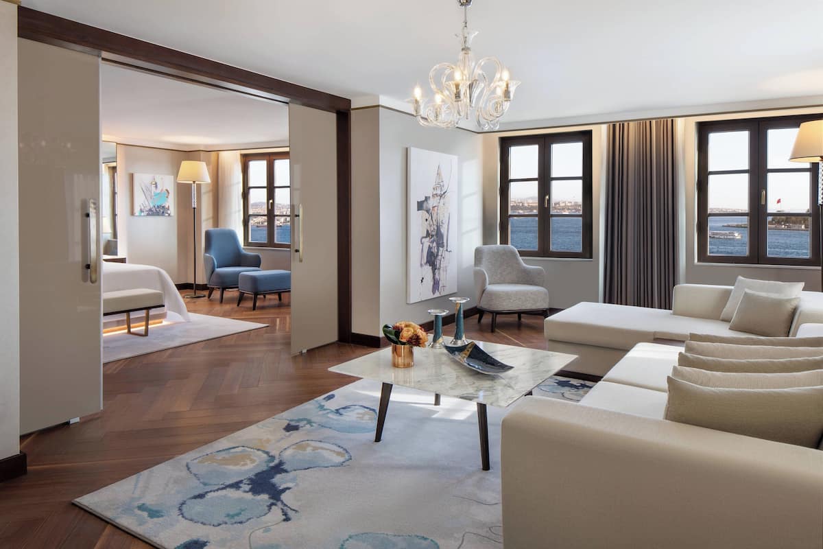 Una habitación de hotel moderna con un ventanal grande, un sofá, una silla, una mesita de centro y una araña.