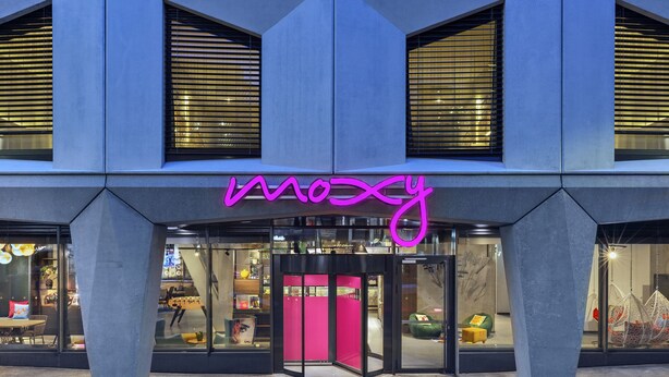 Een modern gebouw met een opvallend neonbord met de tekst 'Moxy' boven de ingang.