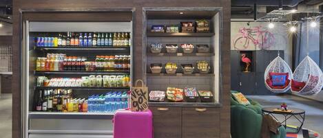 Un expositor de venta al público con diversas bebidas y aperitivos, una maleta rosa y una zona de estar con sillas colgantes.