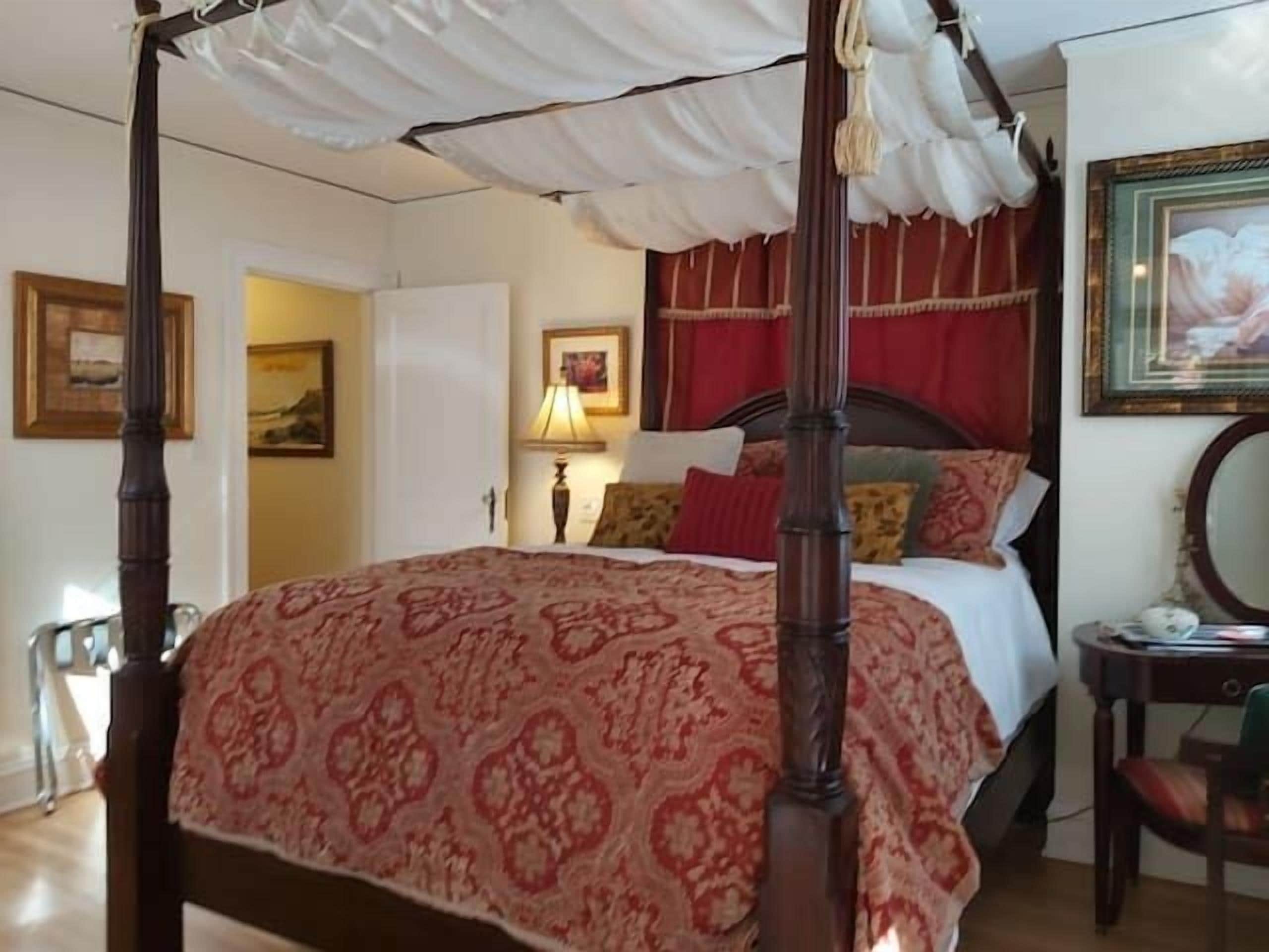 Una cama con dosel, con un edredón rojo estampado, una mesita de noche con lámpara, y un cuadro en la pared.