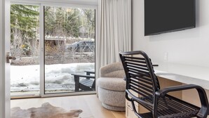 A modern room with a sliding glass door leading to a snowy outdoor area, a comfortable armchair, a black bench, and a flat-screen TV mounted on the wall. - Outpost: Four Seasons I B3 - Recently Renovated! (Teton Village)