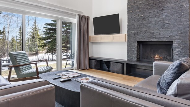A modern living room with a stone fireplace, a flat-screen TV, and a large window overlooking a snowy landscape. - Outpost: Four Seasons I B3 - Recently Renovated! (Teton Village)