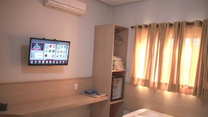 Una habitación de hotel con un televisor montado en la pared, un armario de madera, una cama y una ventana con cortinas.