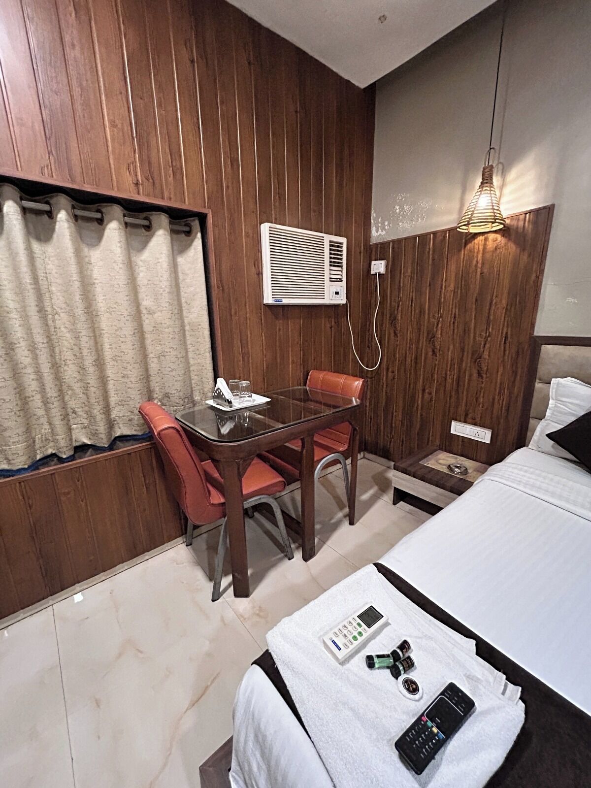 A hotel room with a wooden paneled wall, a single bed with a white comforter, a small table with a glass top and two chairs, a wall-mounted air conditioner, and a bedside table with a lamp.