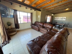 Una sala de estar con un sofá de cuero marrón, una mesa de comedor con sillas, un televisor montado en la pared y una ventana con cortinas.