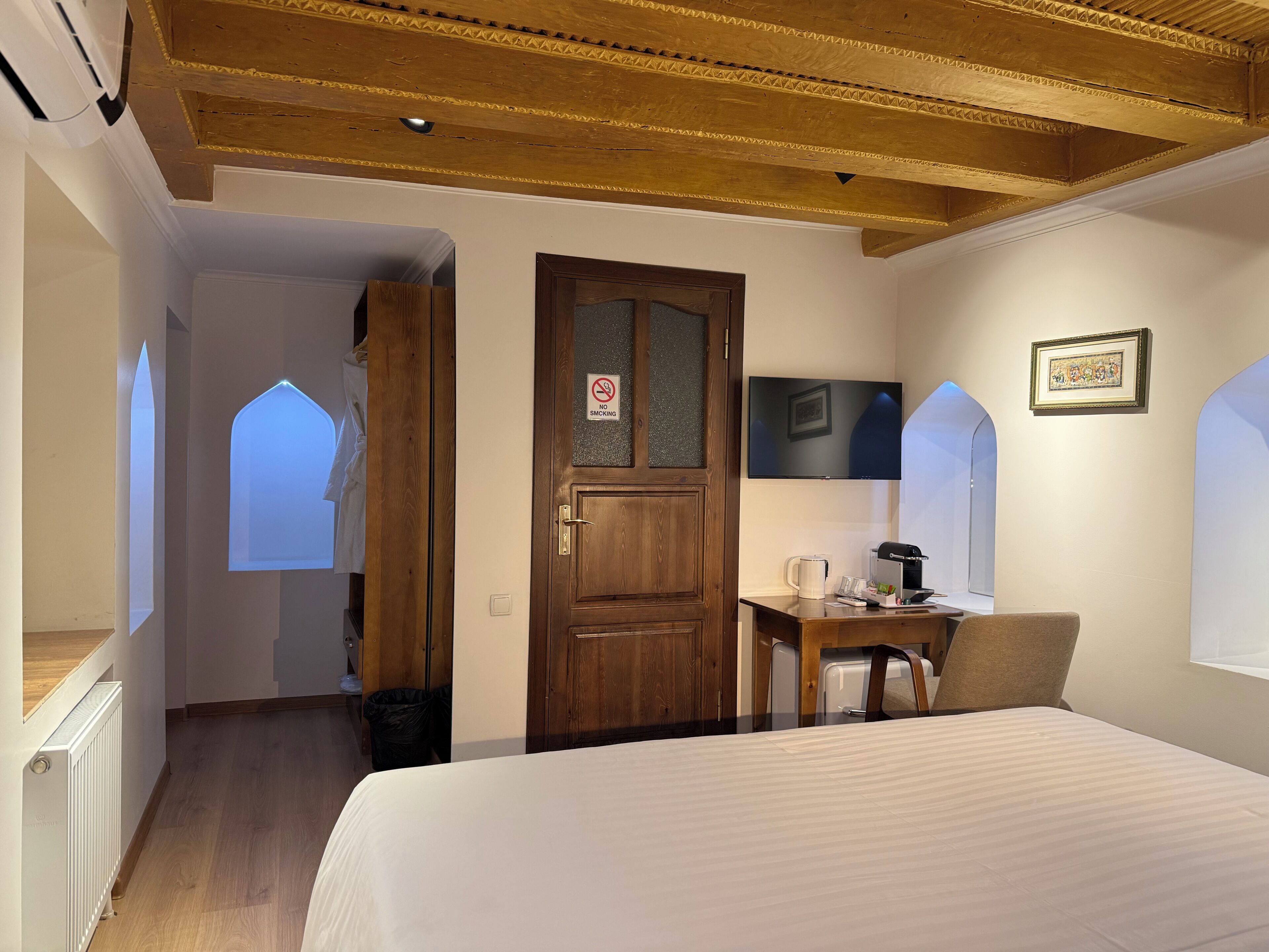 A hotel room with a wooden ceiling, a bed, a desk with a chair, a television, and a 'No Smoking' sign.