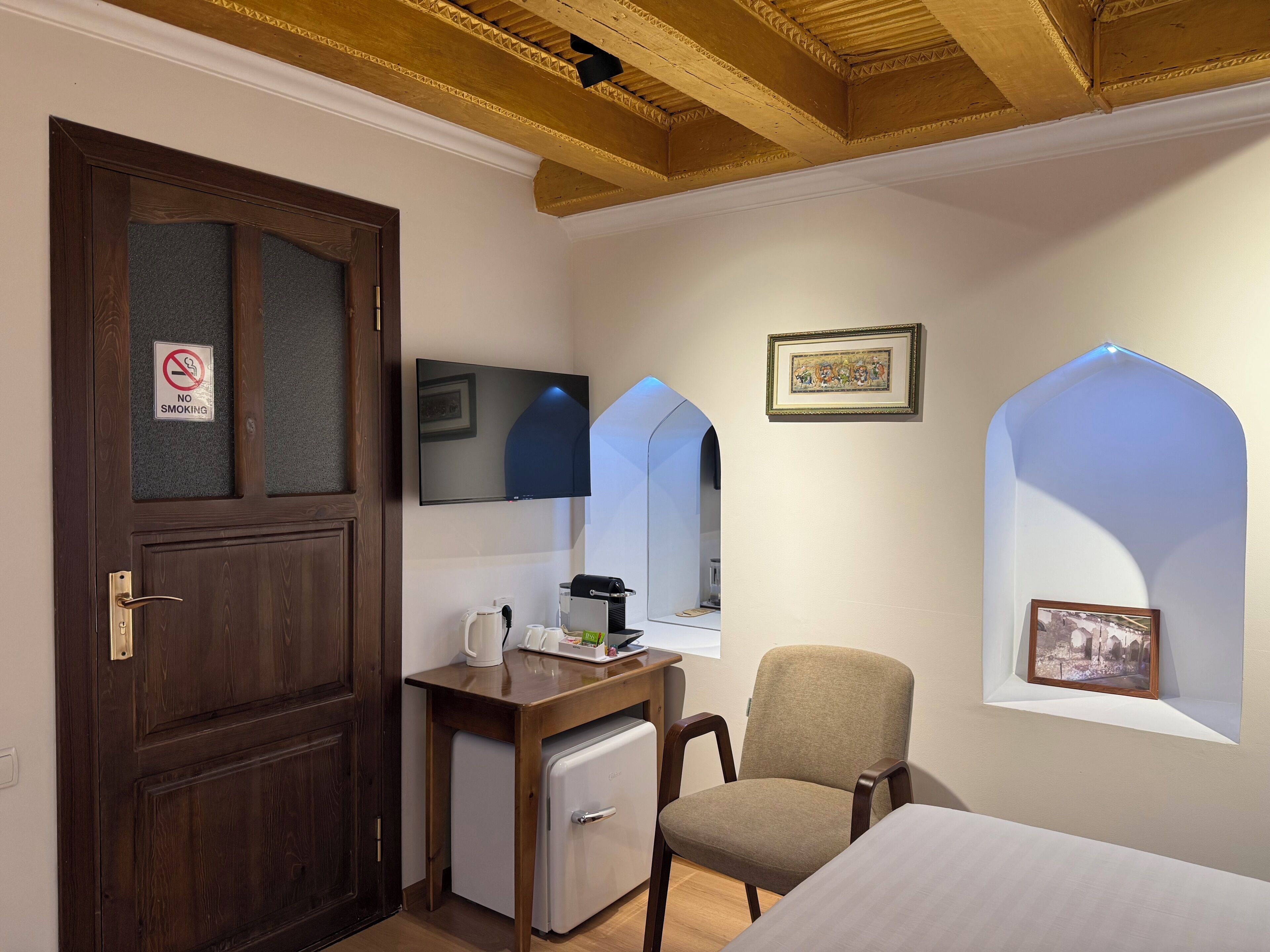 Ein Zimmer mit Holztür, Schreibtisch mit Wasserkocher, Stuhl, Bett, Fernseher und einem Bilderrahmen.