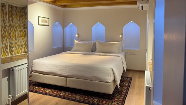 A bedroom with a bed, a radiator, a patterned rug, and arched windows. - Usman Heritage Hotel (Bukhara)
