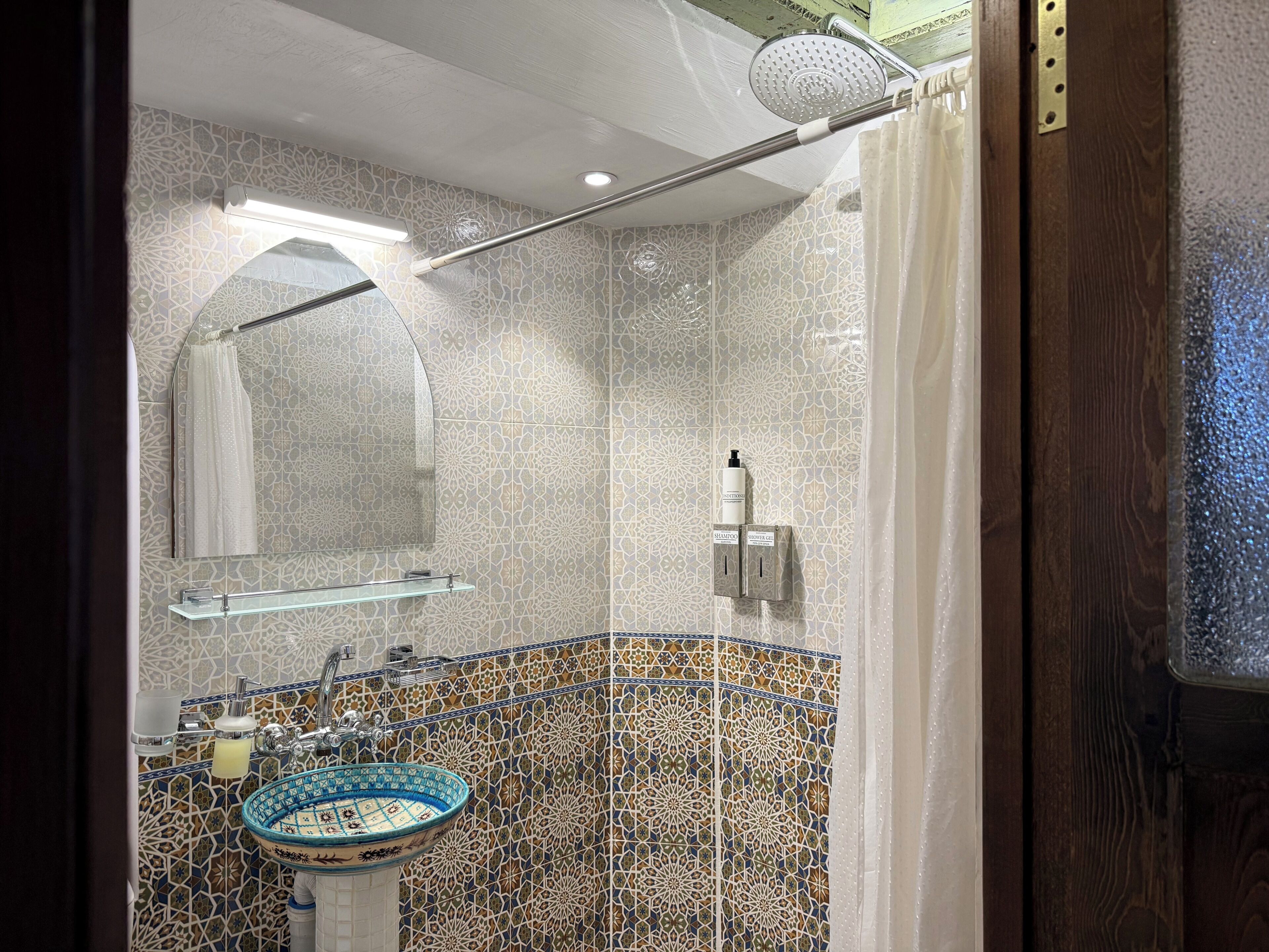 Un baño con pared de azulejos estampados, un lavamanos con lavabo azul y blanco, una zona de ducha con cortina y un accesorio de iluminación en el techo.