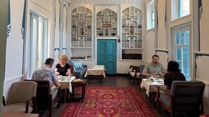 A dining area with a central table surrounded by chairs, a patterned rug, and shelves with dishes and cups. - Usman Heritage Hotel (Bukhara)