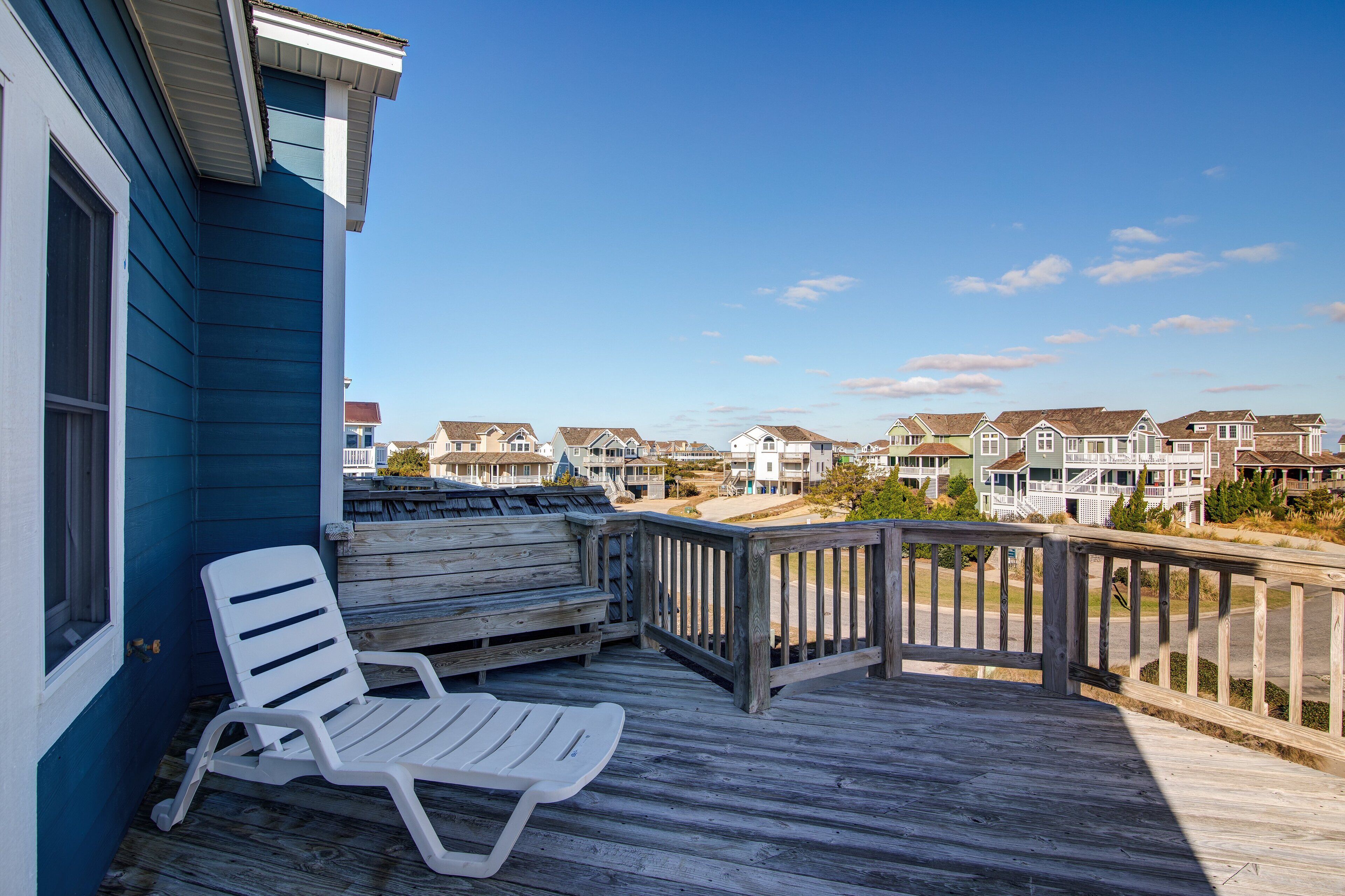 A wooden deck with a white lounge chair, overlooking a row of houses with a clear blue sky.