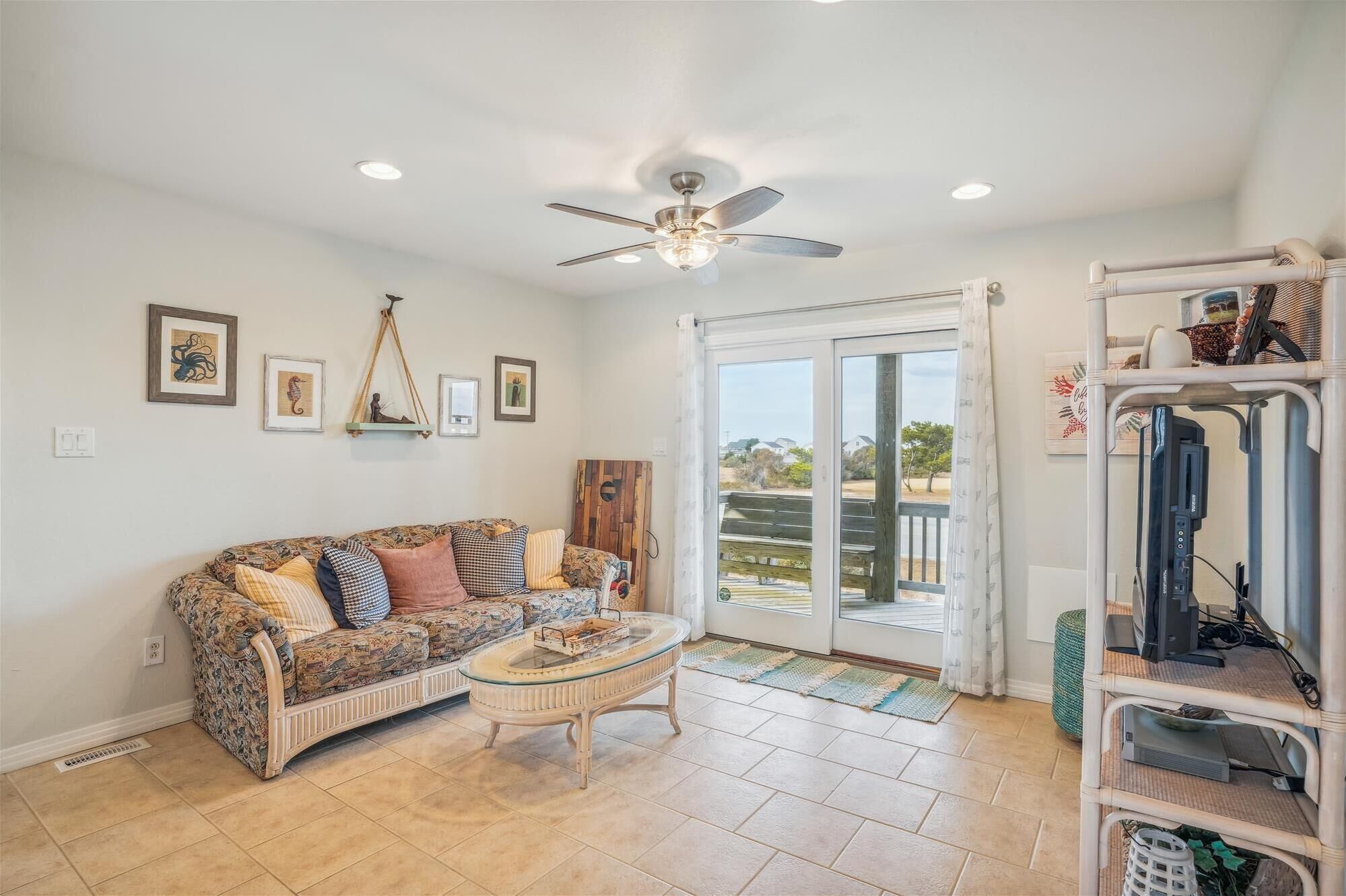 A living room with a patterned sofa, a glass coffee table, a ceiling fan, and a sliding glass door leading to a balcony.