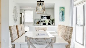 Un coin repas avec une table blanche et des chaises en osier, un luminaire suspendu et une cuisine en arrière-plan.