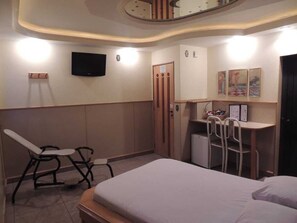 Quarto de hotel com cama, escrivaninha, cadeira, televisão e pequena geladeira.